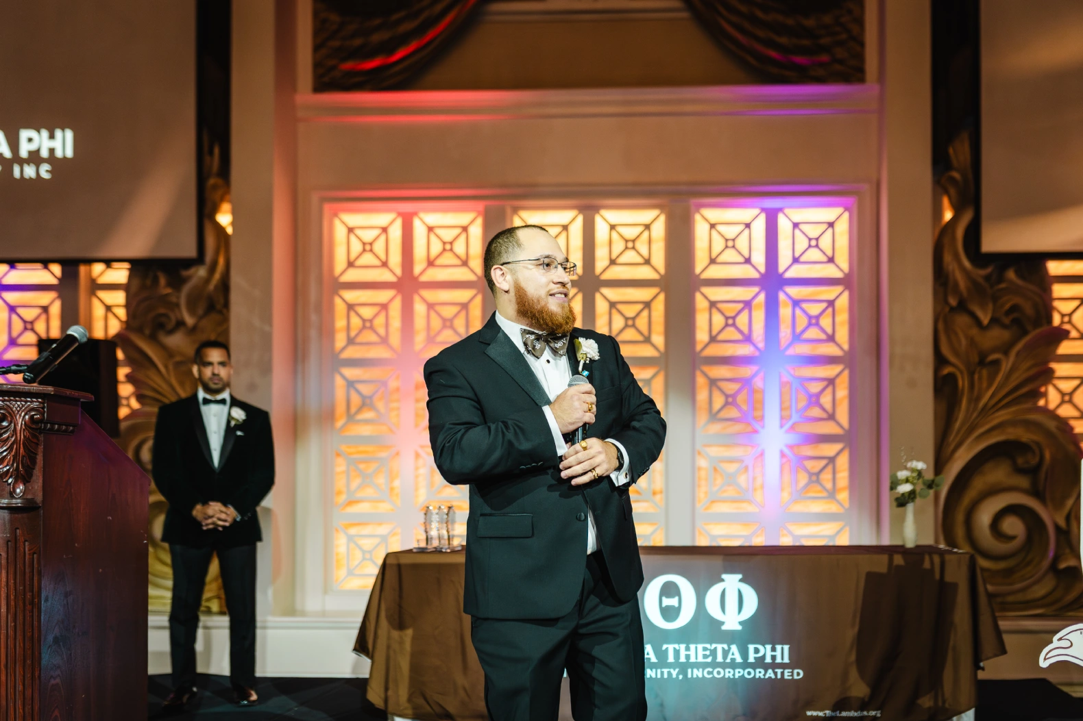 Man with a red beard in a black suit holding a microphone at a Lambda Theta Phi fraternity event with colorful lit background.