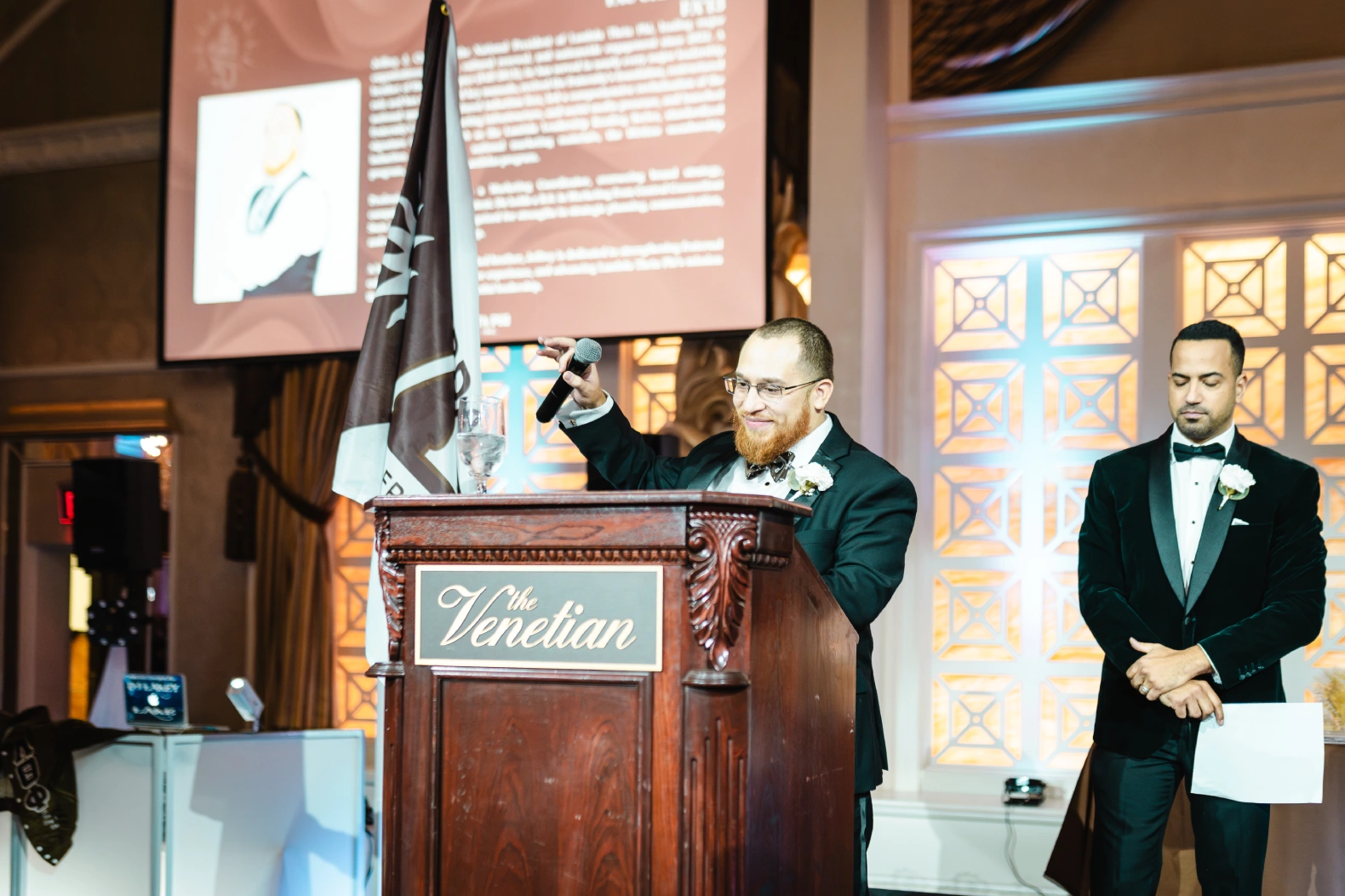 Two men in tuxedos at a podium with a microphone and a flag, one speaking and the other holding a paper.