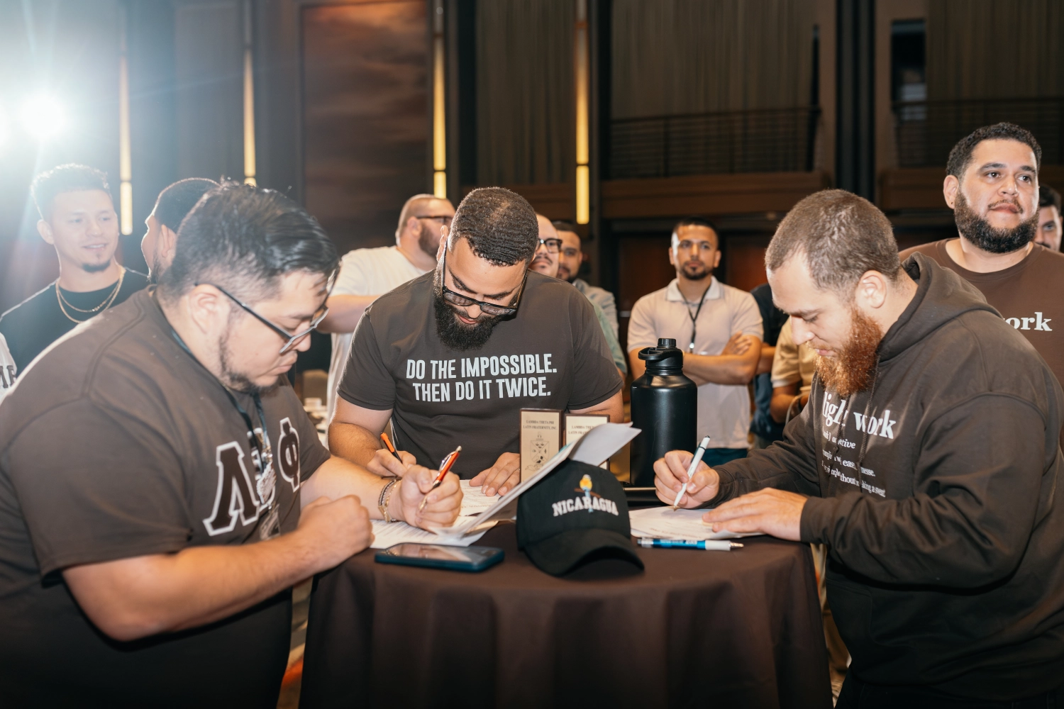 Three men with beards writing on papers at a round table with a Nicaragua hat and a large black water bottle, surrounded by people in a well-lit room.