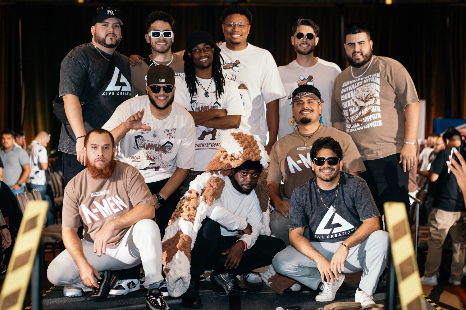Group of eleven men posing together indoors, some wearing sunglasses and casual clothing with printed designs.