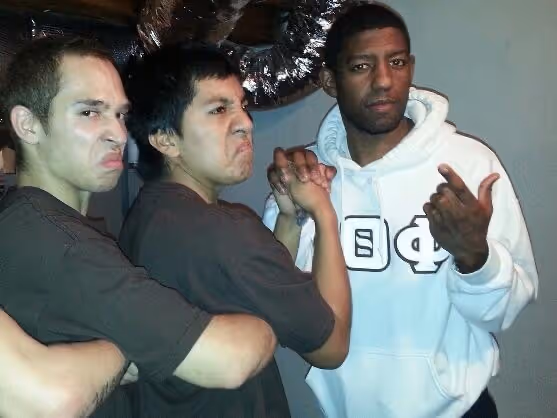 Three young men posing together against a background of ductwork in a basement or industrial setting, two with crossed arms and serious expressions, and one wearing a white hoodie with Greek letters.