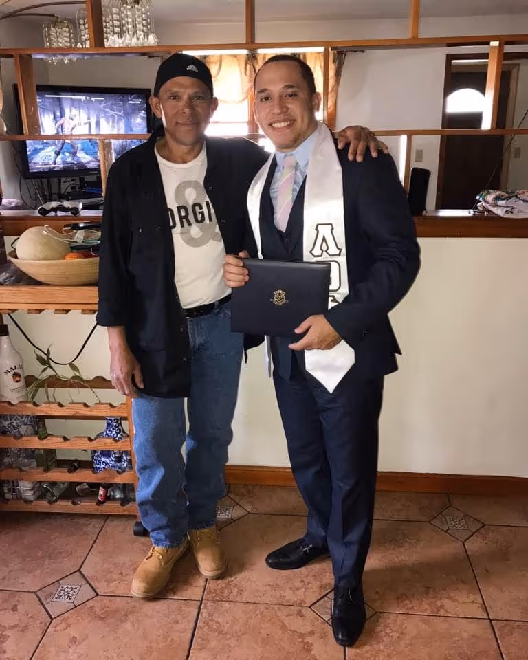 Two men standing indoors; one dressed casually in jeans and a cap, the other in a suit with a white graduation stole holding a diploma cover.