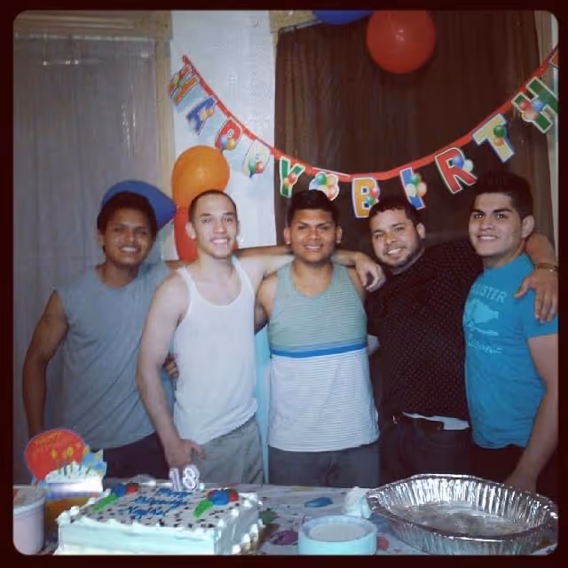 Five young men smiling together at an 18th birthday party with a decorated cake and balloons in the background.