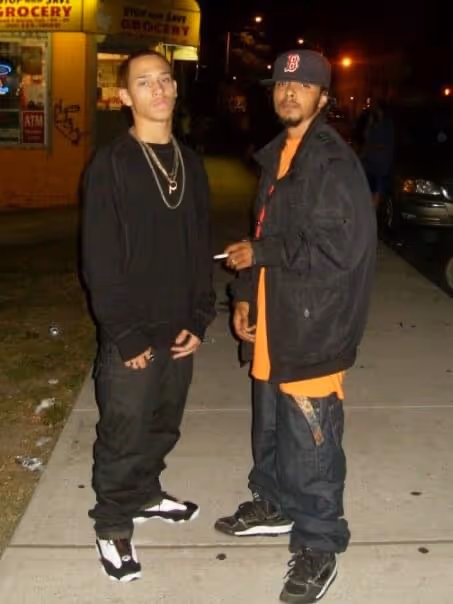 Two young men standing on a sidewalk at night, one wearing a black outfit with white and black sneakers, and the other in a black jacket, orange shirt, and baseball cap holding a cigarette.