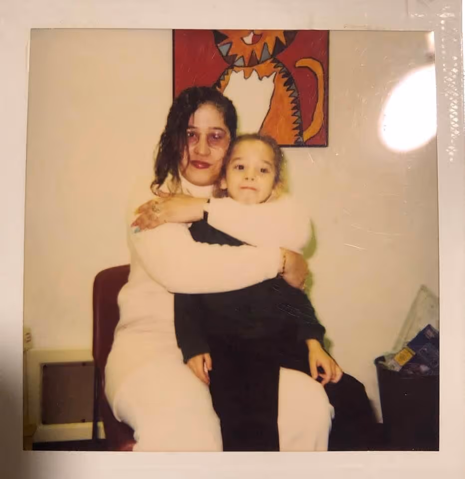 Woman in white outfit hugging a child dressed in black sitting on her lap, with a colorful cat painting on the wall behind them.