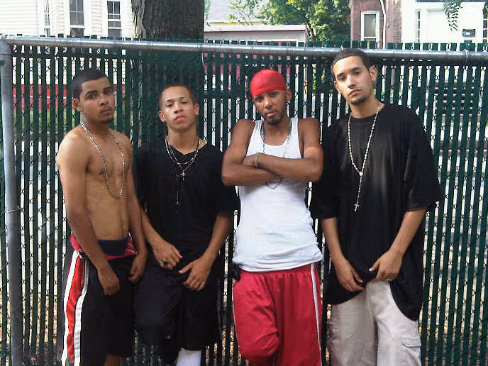 Four young men standing in front of a green metal fence with trees and houses in the background, dressed in casual streetwear including tank tops, t-shirts, and shorts.