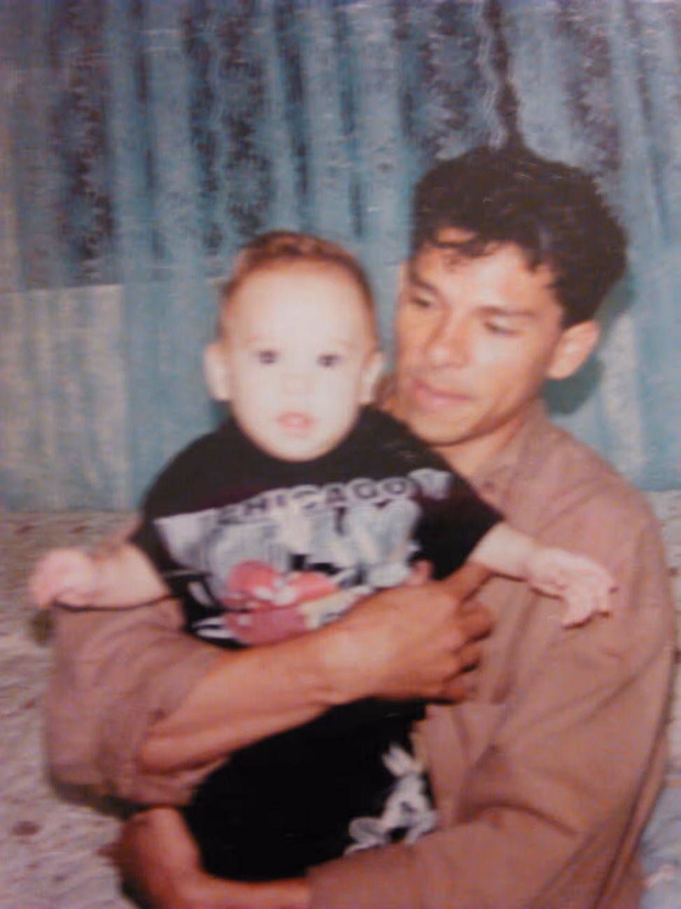 A man in a brown shirt holding a baby wearing a black Chicago Bulls shirt in front of a blue curtain.