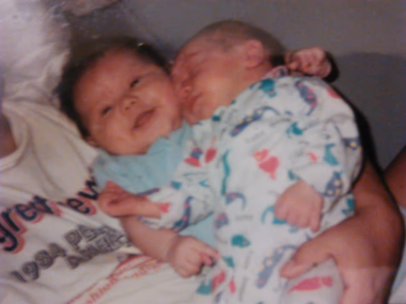 Two babies in printed onesies, one smiling and the other sleeping, being held by an adult wearing a white shirt.