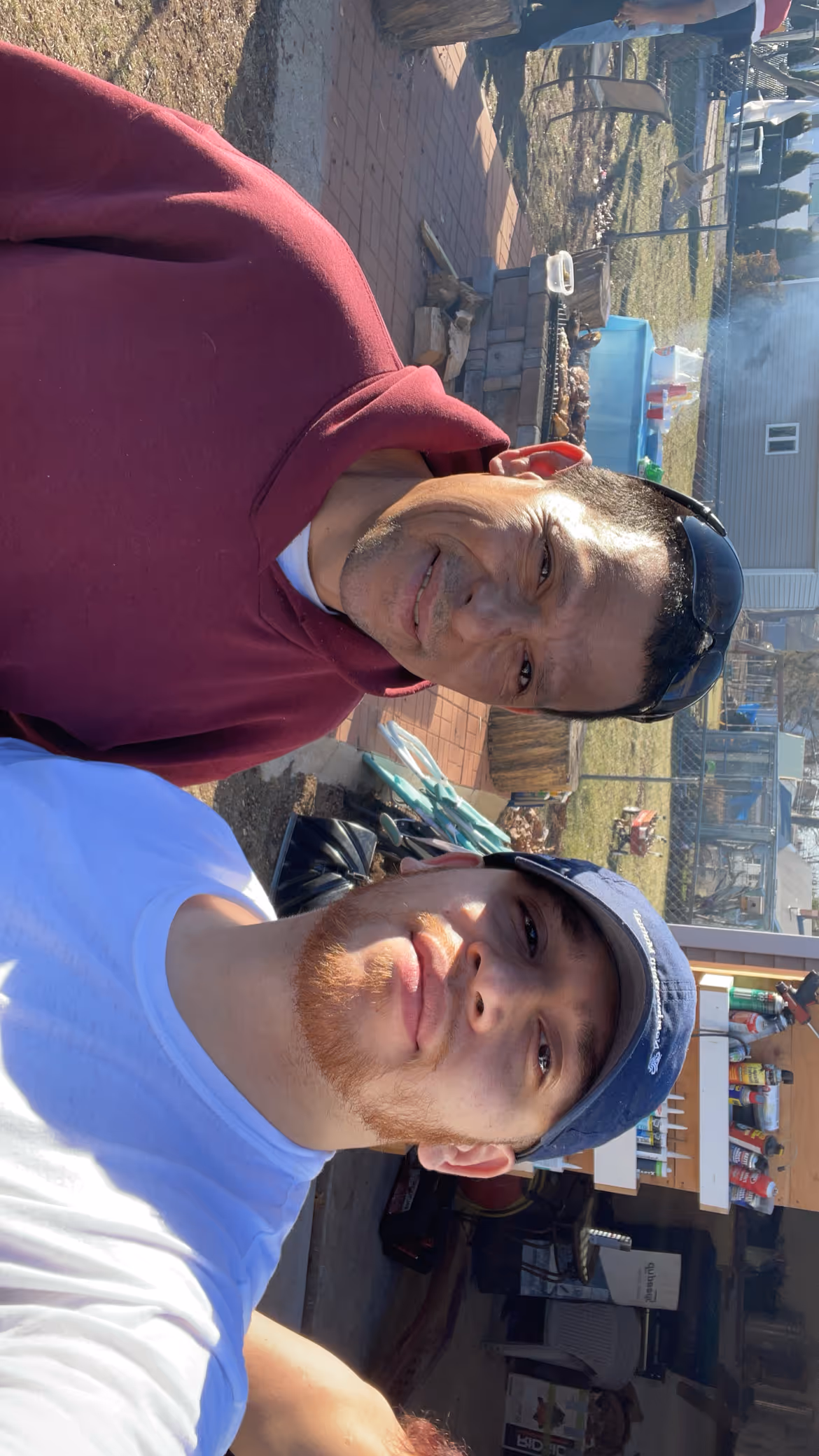 Two men outdoors posing for a selfie; one wears a maroon hoodie and sunglasses on his head, the other has a reddish beard and a navy blue cap.