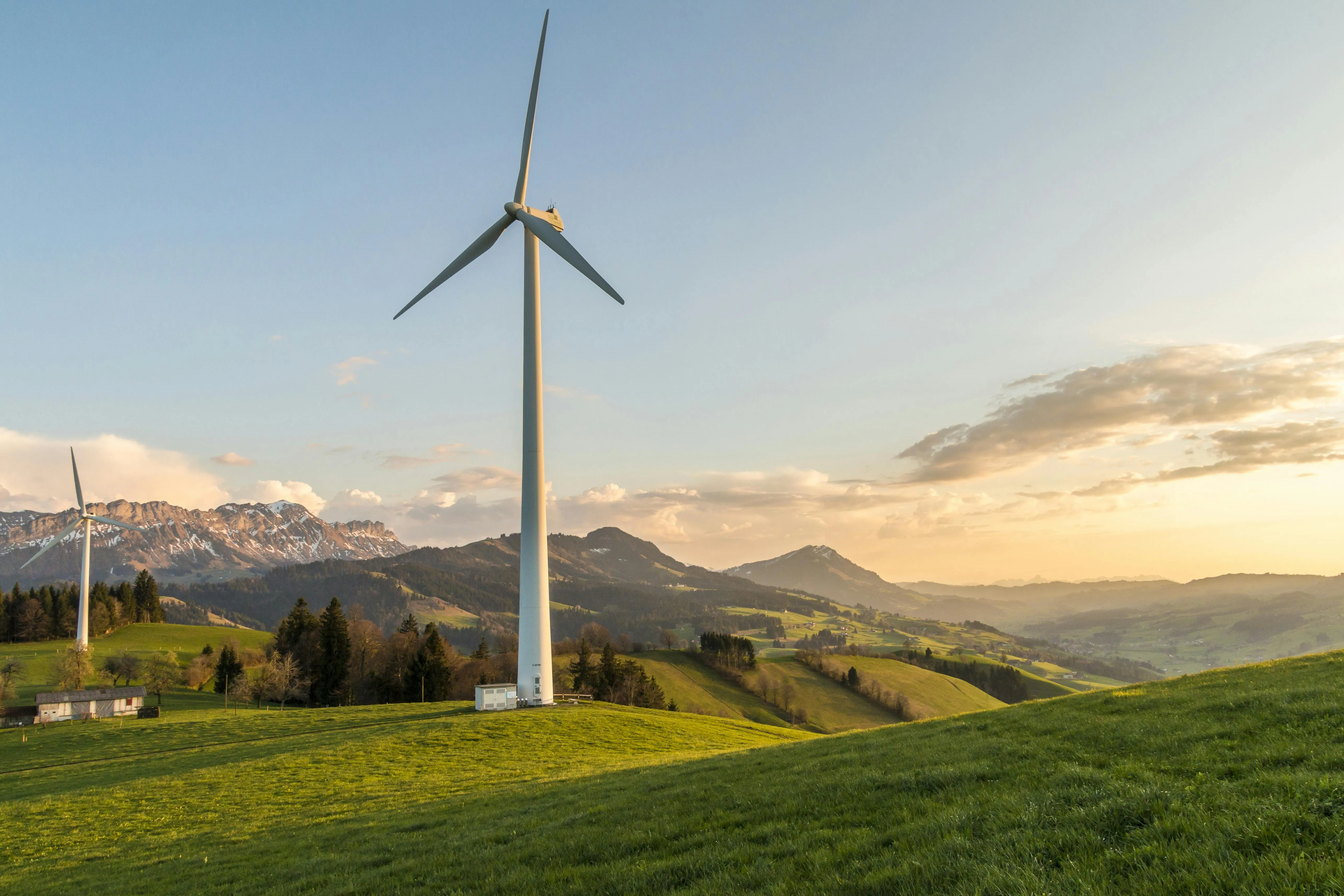 Wind turbines on green rolling hills with mountains in the background during sunset.