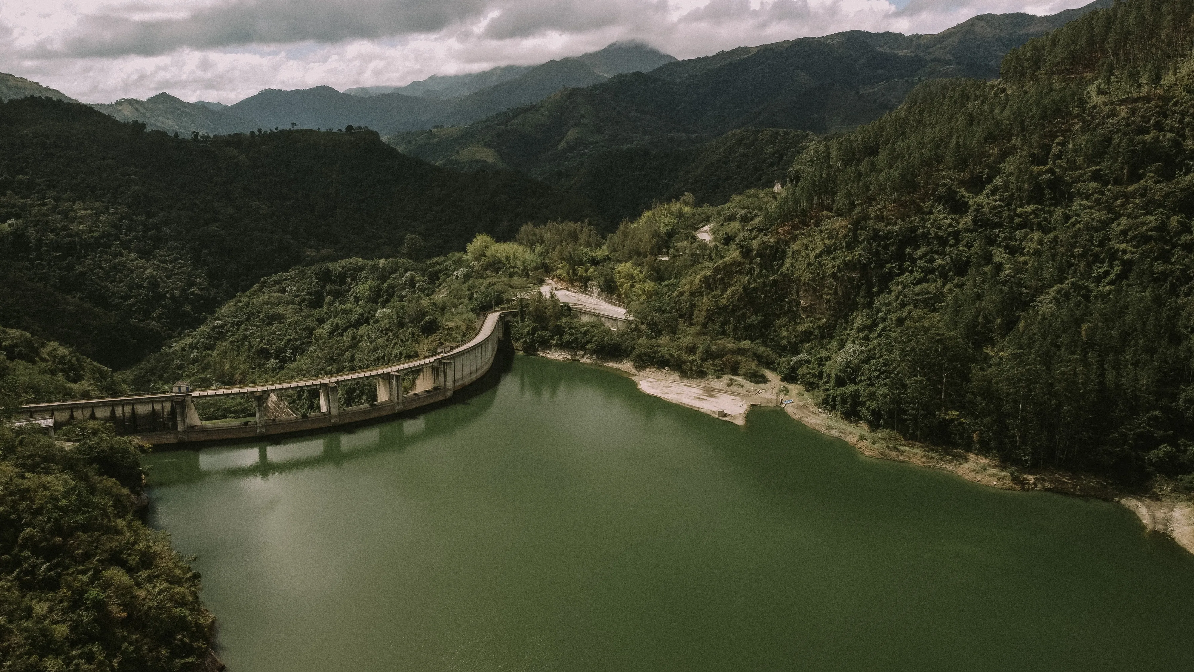 Dam reservoir surrounded by dense green forested mountains under a cloudy sky.