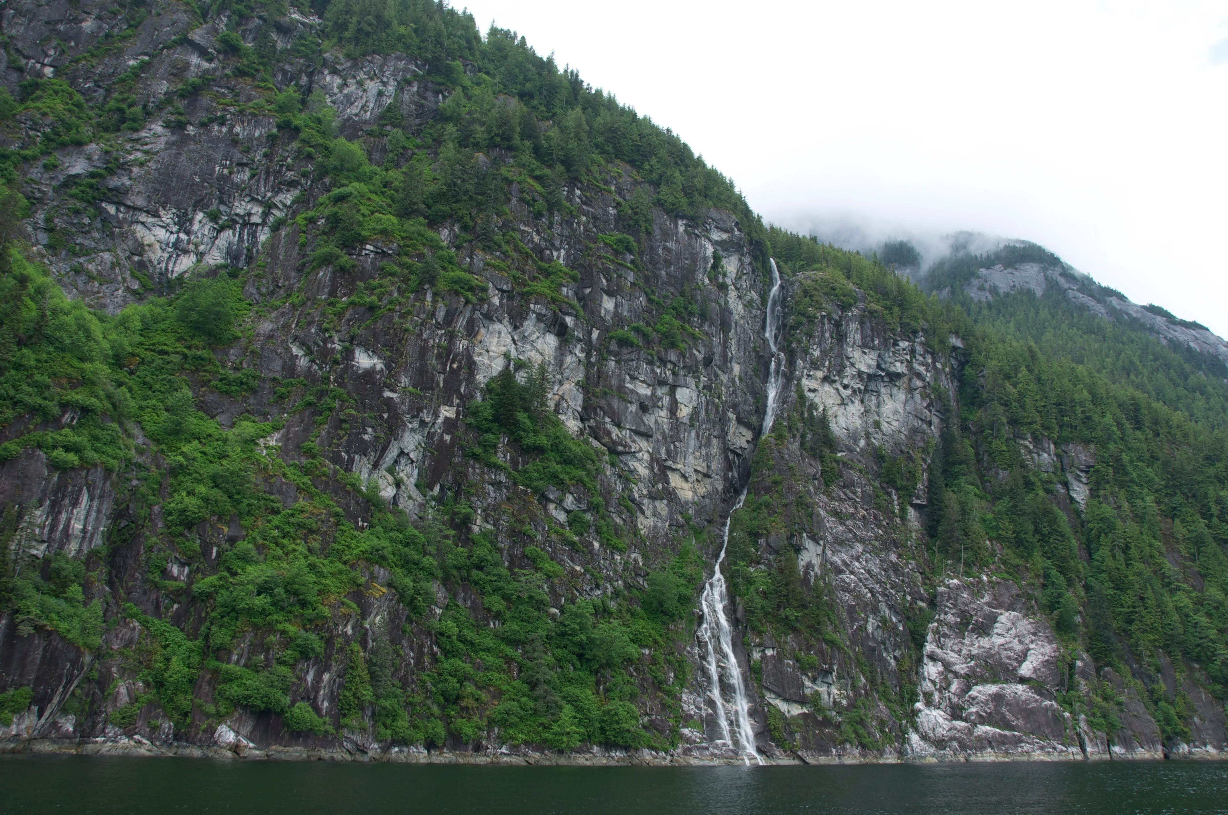 Tall, narrow waterfall cascading down a rocky, forested mountain into a body of water.