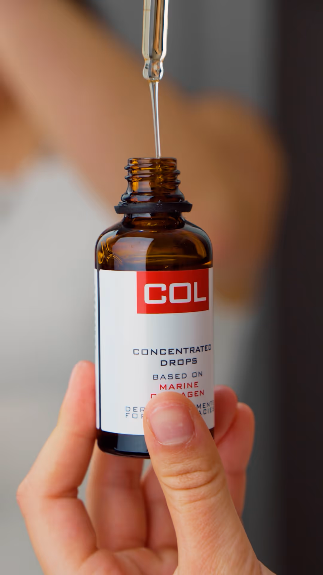 Hand holding a brown glass bottle labeled COL with a dropper releasing a clear liquid into it.
