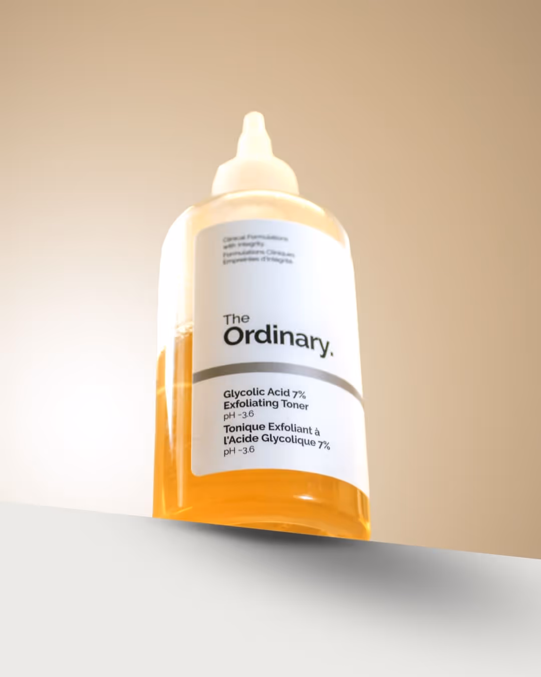 Bottle of The Ordinary Glycolic Acid 7% Exfoliating Toner with yellow liquid inside on a white surface and beige background.