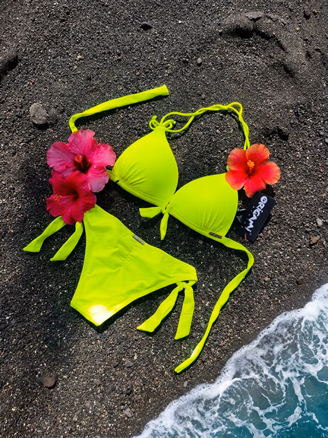 Neon yellow bikini with pink and red flowers on black sand near ocean waves.