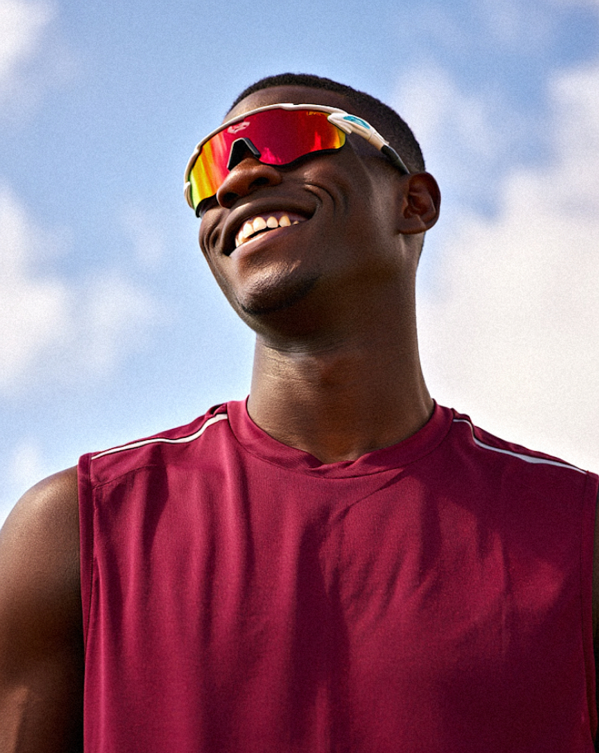 Smiling man wearing red sports sunglasses and a sleeveless maroon shirt against a blue sky.