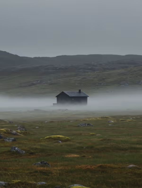 Small dark cabin surrounded by green mossy fields with low fog and rolling hills in the background under a gray sky.