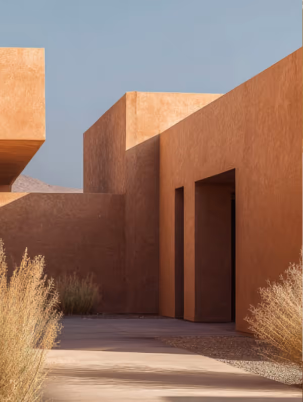 Minimalist desert building with angular tan stucco walls, narrow doorways, and dry shrubbery under clear sky.