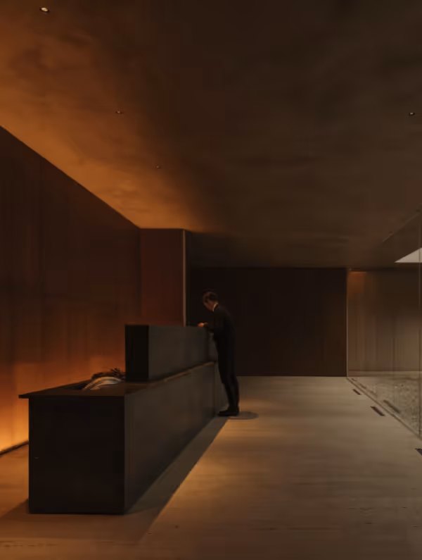 Person standing at a minimalist dark reception desk in a warmly lit modern interior space with wooden and concrete surfaces.