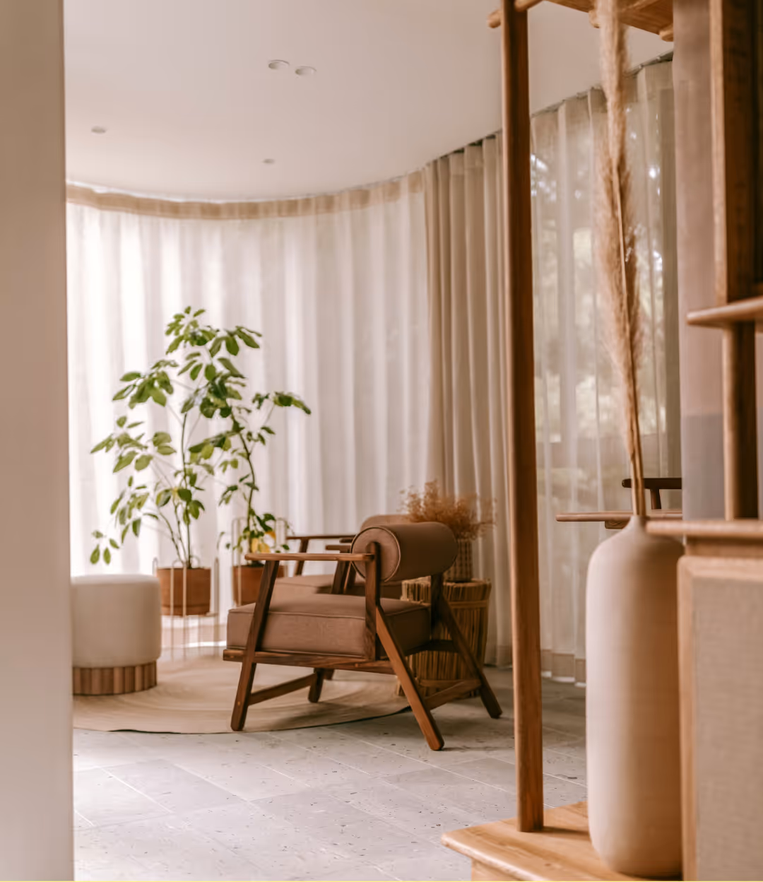 Cozy modern corner with a brown cushioned wooden chair, green potted plants, sheer beige curtains, and minimalist decor.
