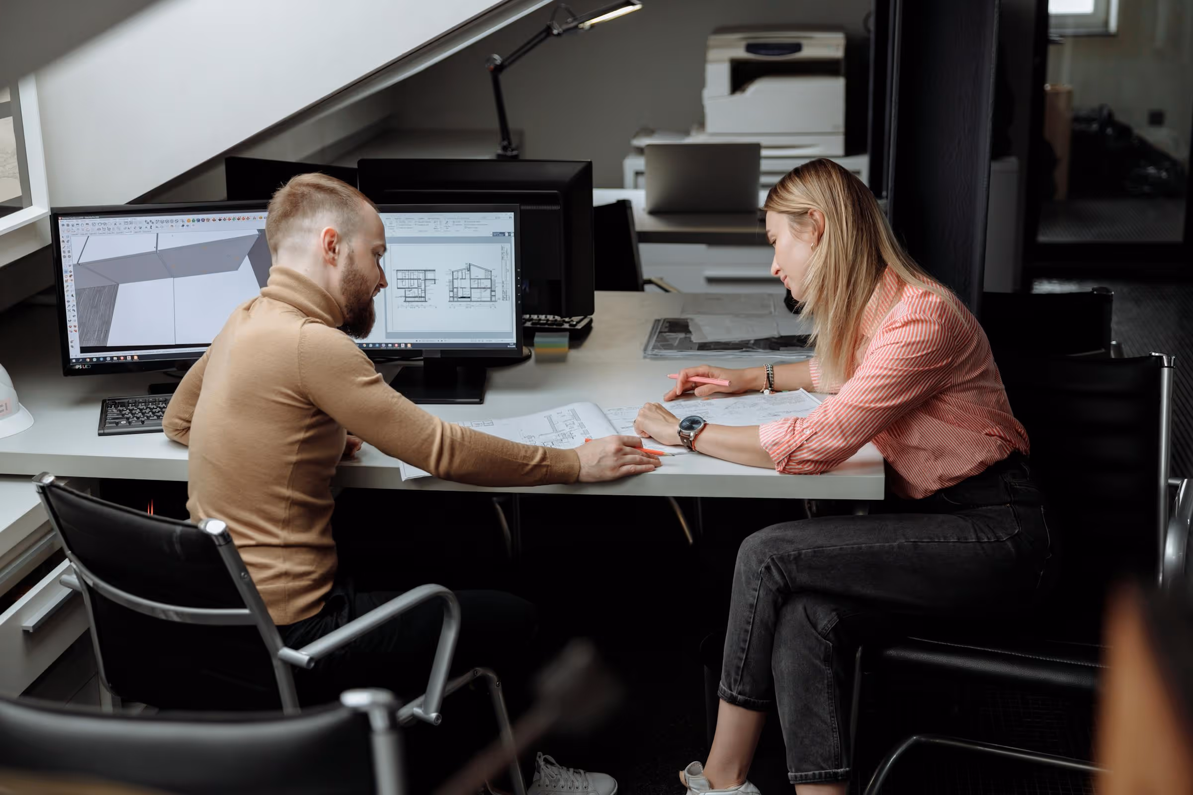 Two architects discussing building plans at a desk with computer monitors displaying architectural designs.