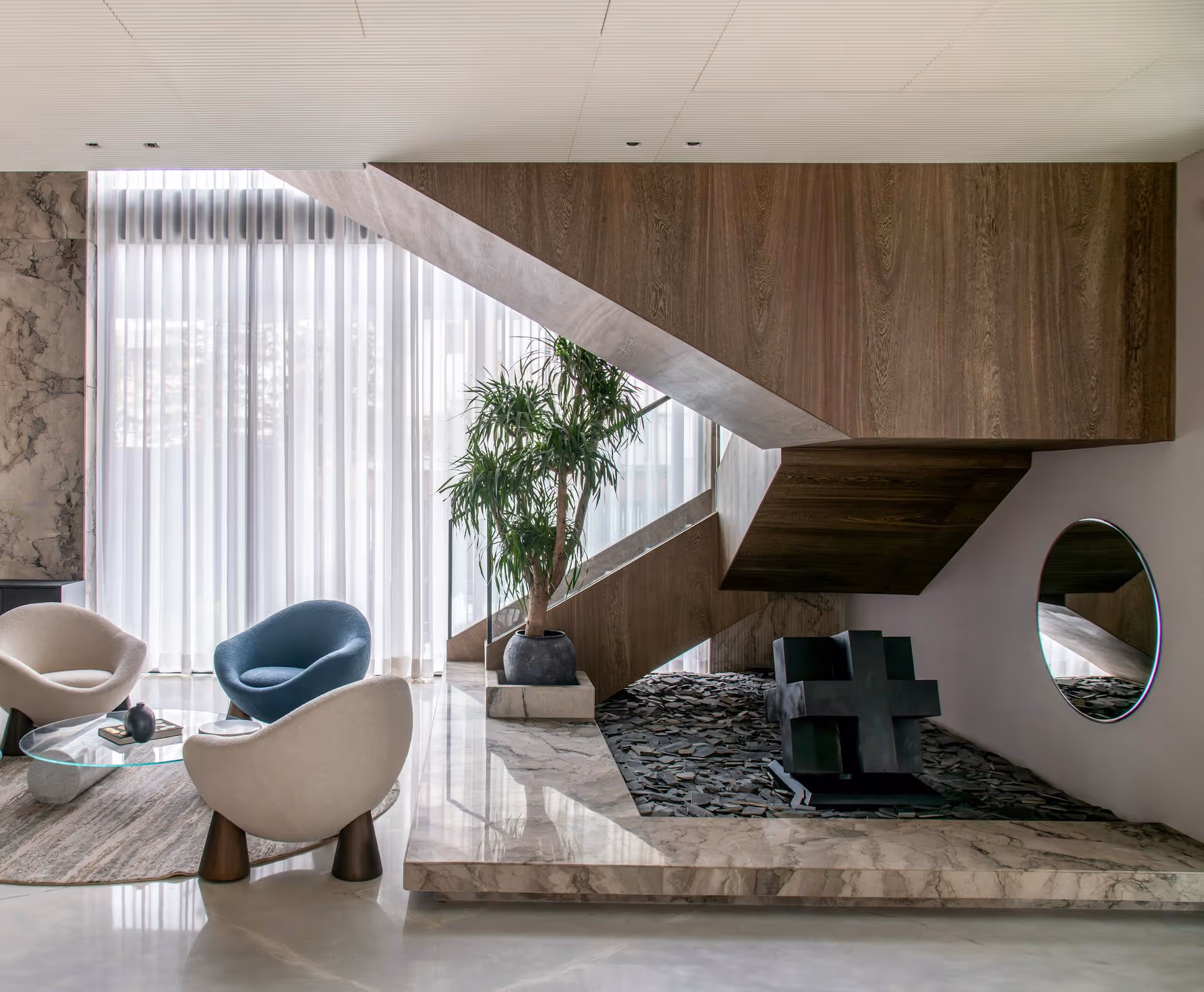 Modern living room with three stylish chairs, a glass coffee table, a potted plant, wooden staircase, and a circular wall mirror.