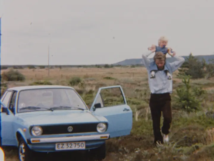 Man carrying a child on his shoulders walking beside an open-door blue vintage car in a rural landscape.