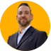 Portrait of a smiling man with short hair and beard wearing a dark blazer over a white shirt against a yellow background.