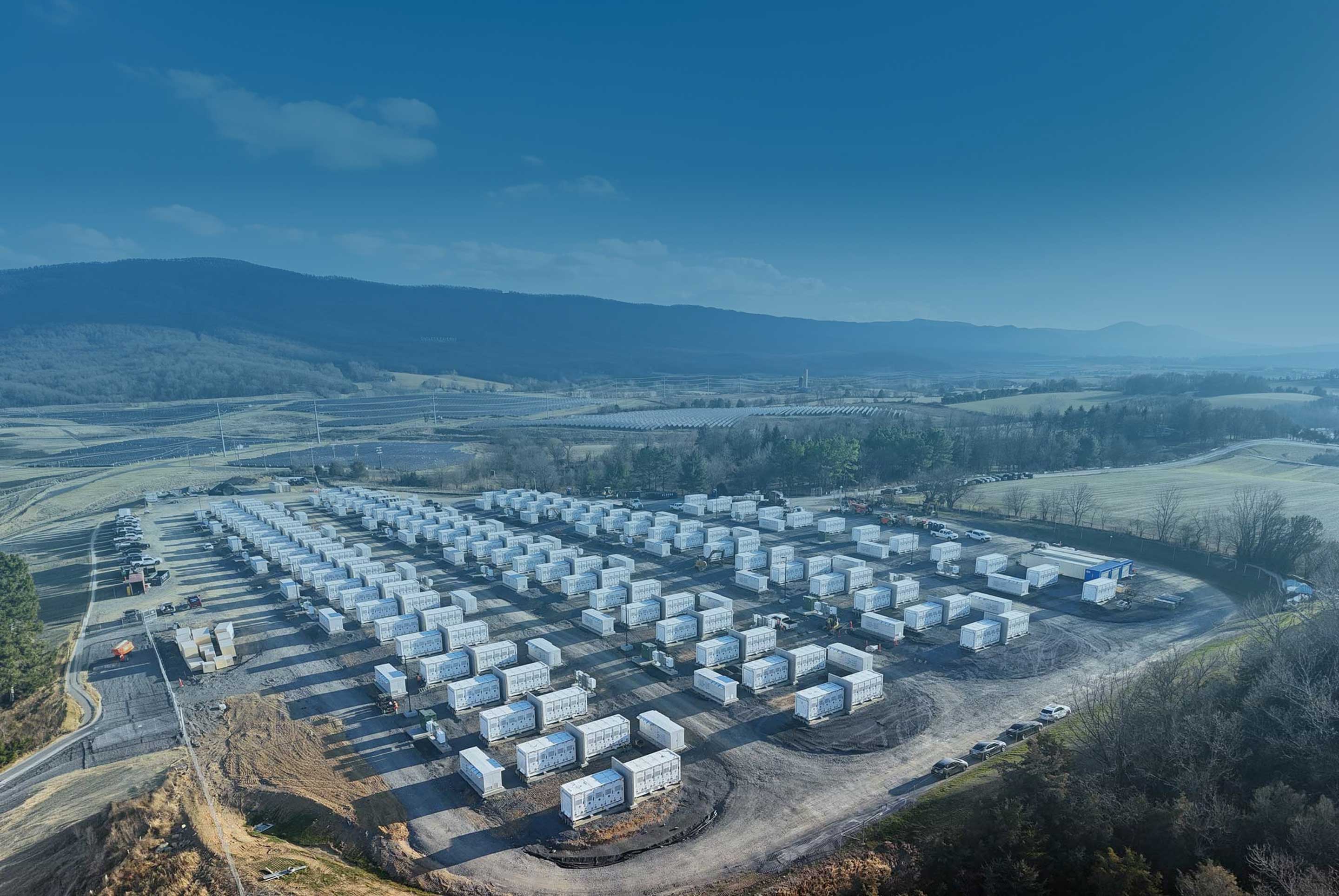 Aerial view of a large solar battery storage facility with rows of white battery storage units on a cleared plot surrounded by fields and hills.
