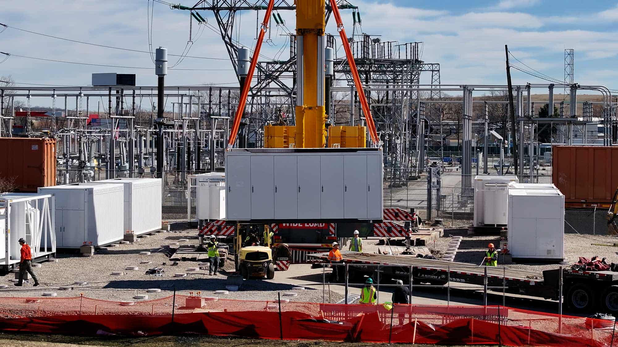 Construction workers operating a crane to lift a large electrical container in a power substation.