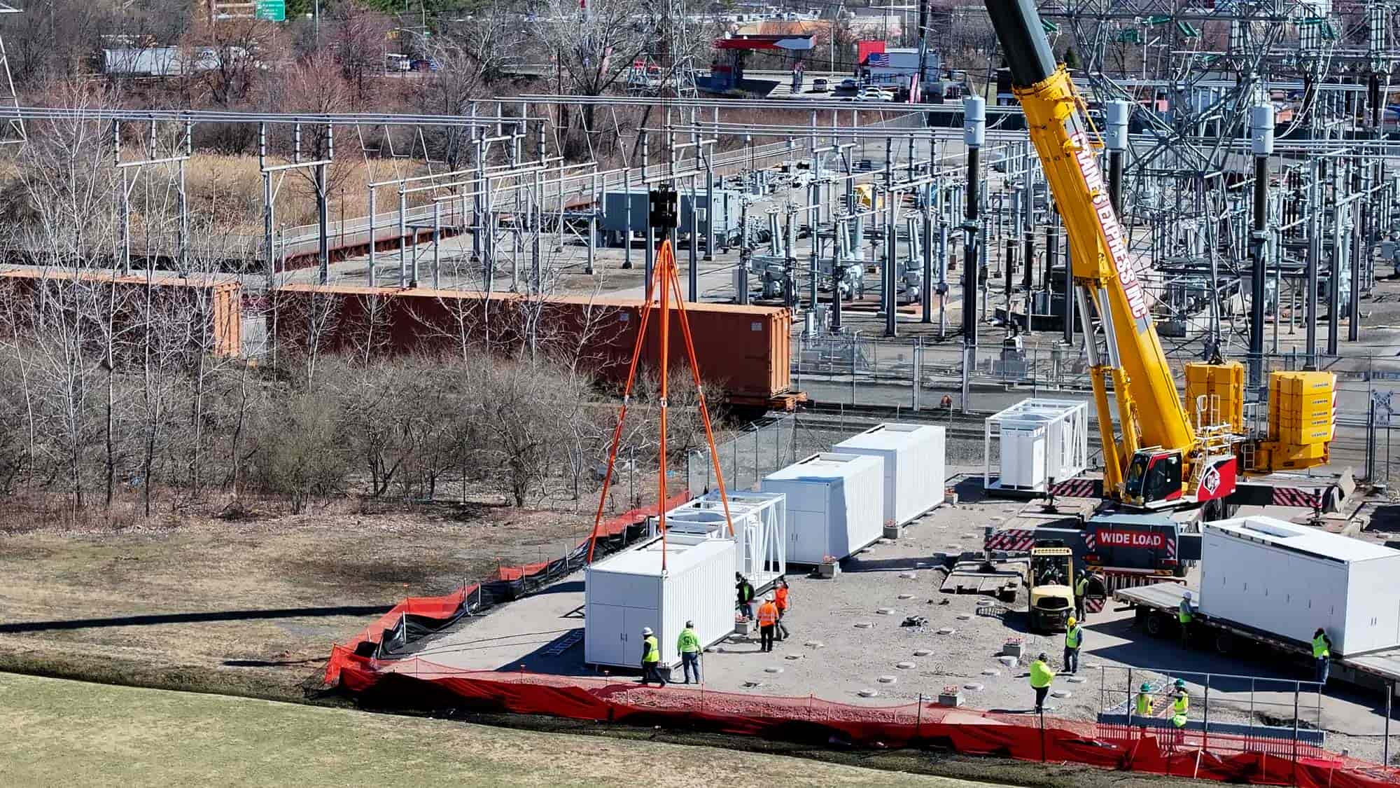Construction workers operating a crane to lift a large electrical container in a power substation.