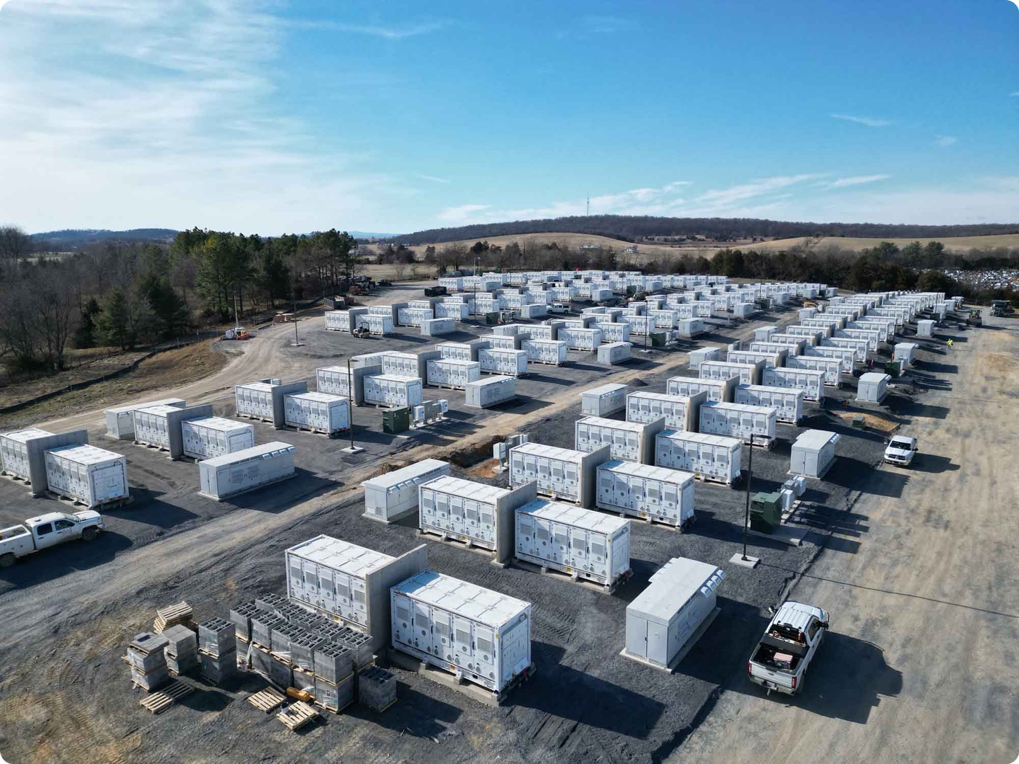 Aerial view of an industrial yard with multiple rows of large white electrical units, several white trucks, and pallets, surrounded by trees and hills under a partly cloudy sky.