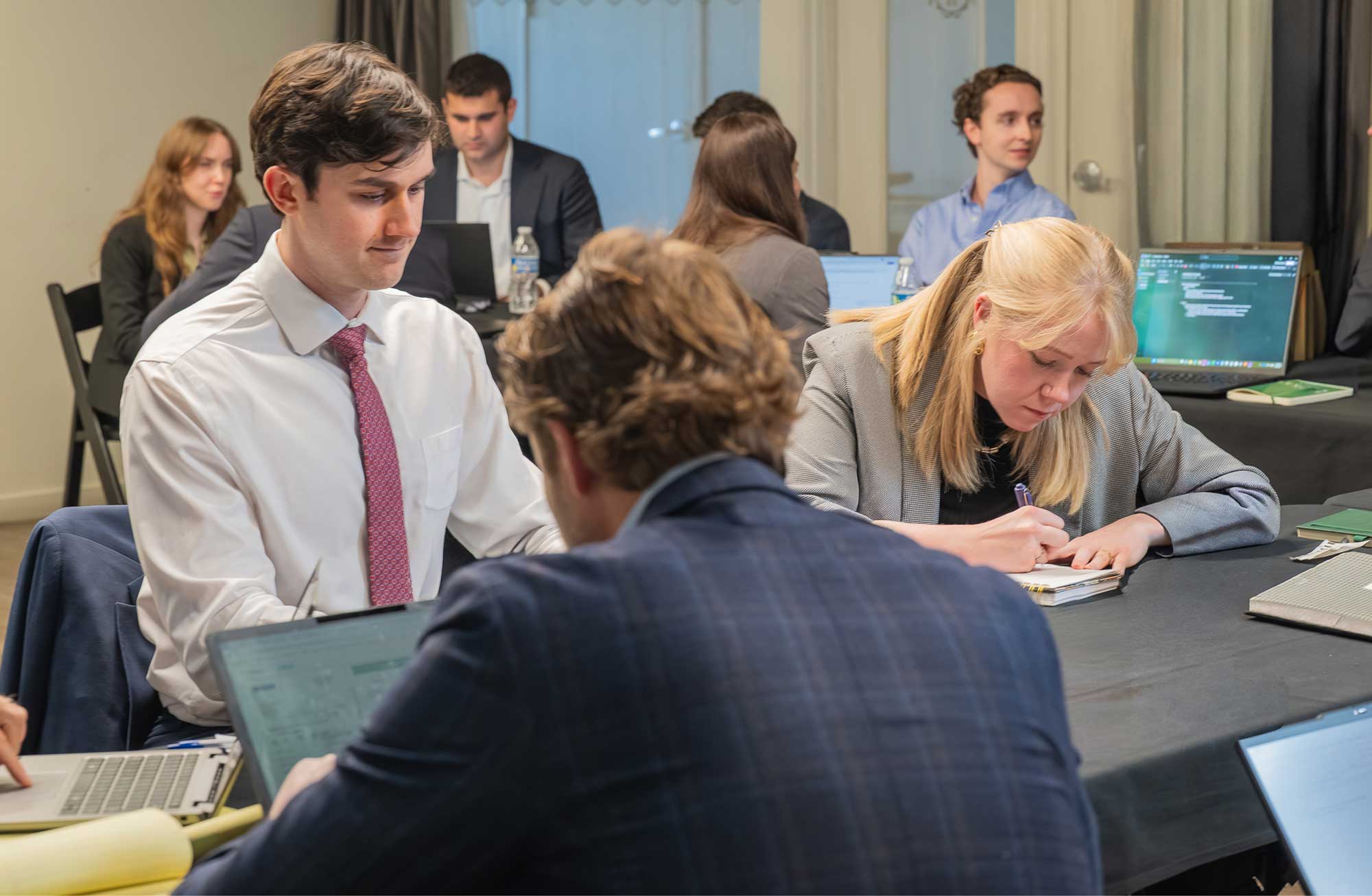 Group of young professionals working together at a table with laptops and notebooks in a collaborative office setting.