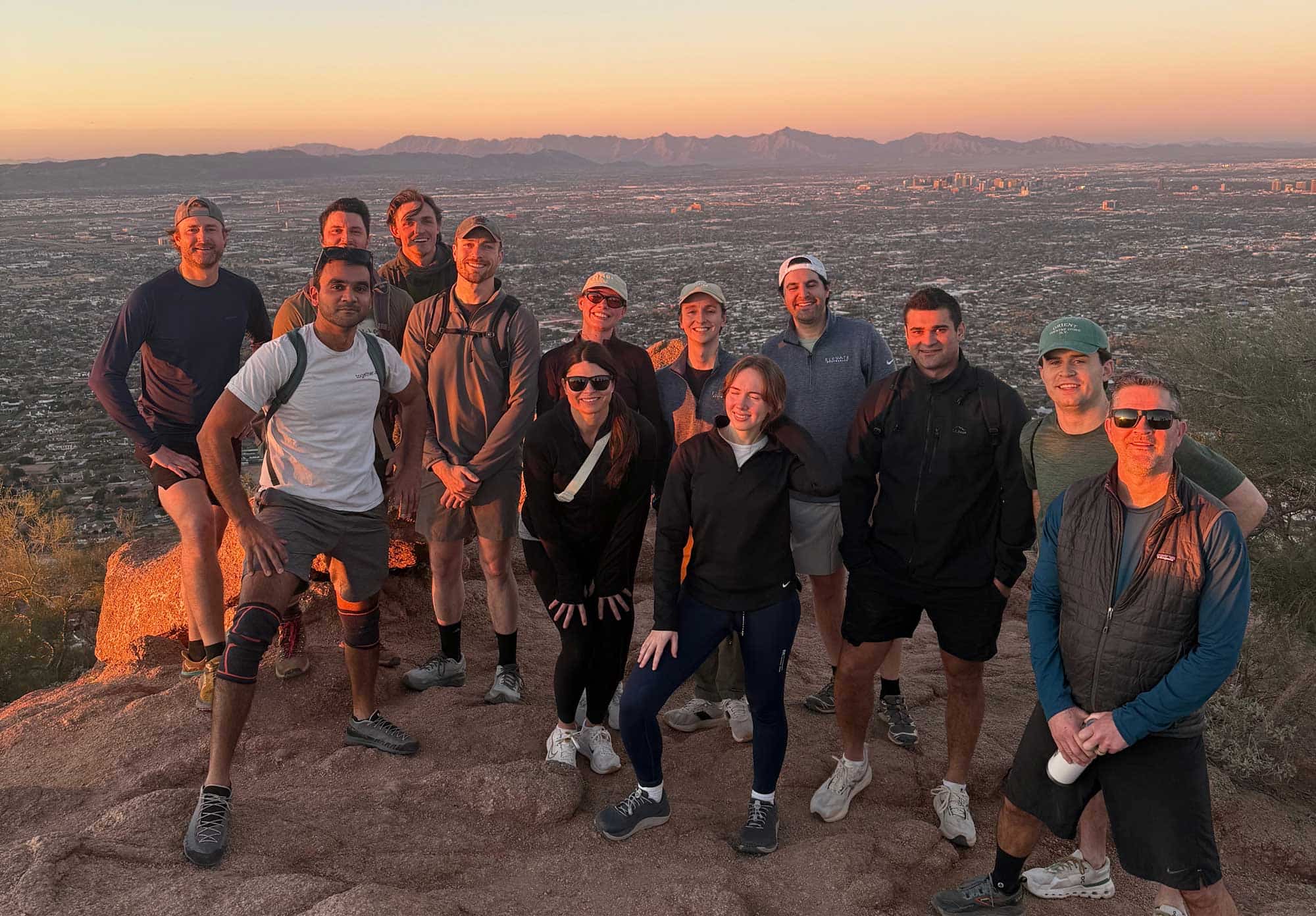 Group of thirteen people posing on a mountain during sunset with a city and mountains in the background.