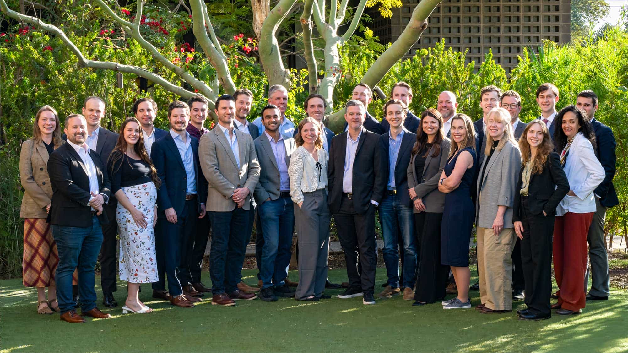 Group of 25 professionals standing outdoors on grass with trees and greenery in the background, smiling at the camera.