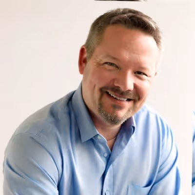 Smiling man with short brown hair and a goatee wearing a light blue button-up shirt against a plain background.