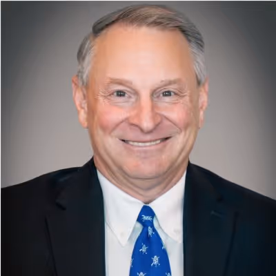 Smiling middle-aged man in a dark suit, white shirt, and blue patterned tie against a neutral background.