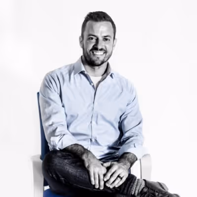 Smiling man with short dark hair wearing a light blue shirt and dark jeans sitting cross-legged on a chair against a white background.