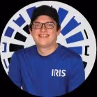 Smiling man wearing glasses, a black cap, and a blue IRIS t-shirt against a blue and white circular background.