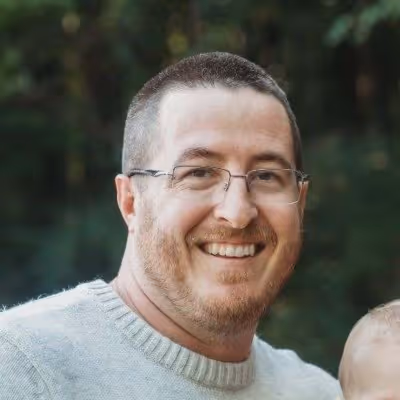 Smiling man with short hair, glasses, and a gray sweater against a blurred green outdoor background.