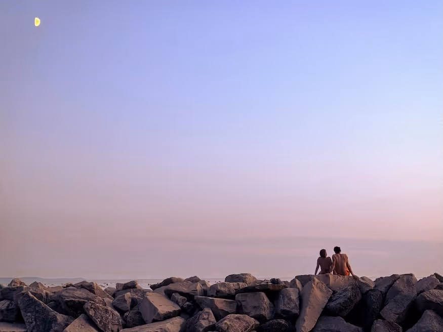 Two people sitting on large rocks facing the calm ocean under a pastel purple and blue sky with a visible crescent moon.