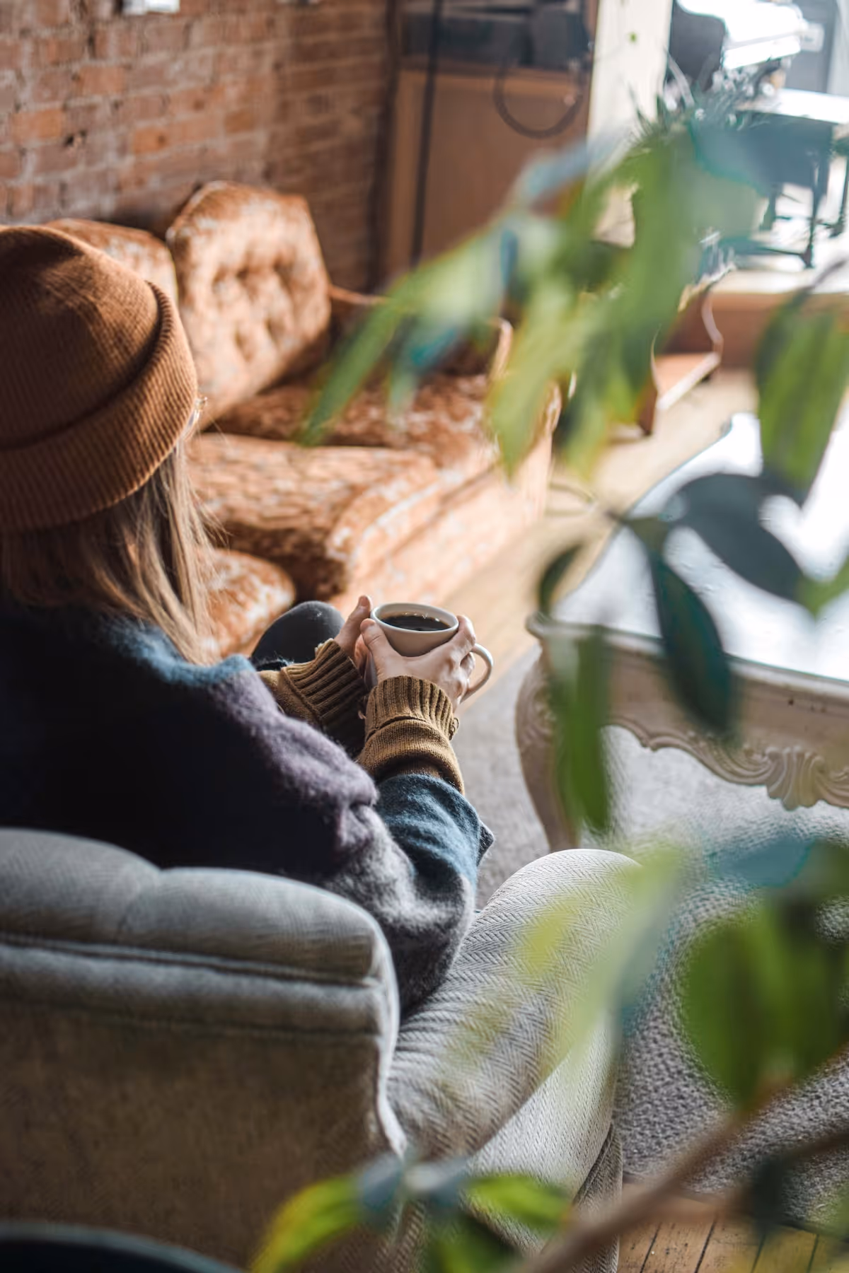Person wearing a brown beanie and sweater sitting on a beige armchair holding a white cup of coffee in a cozy living room with a vintage sofa and a wooden table.