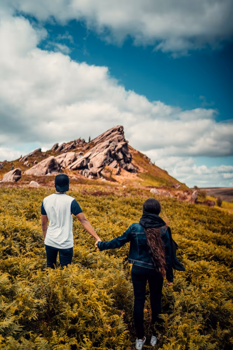 Couple holding hands walking through green ferns towards rocky hill under a partly cloudy sky.