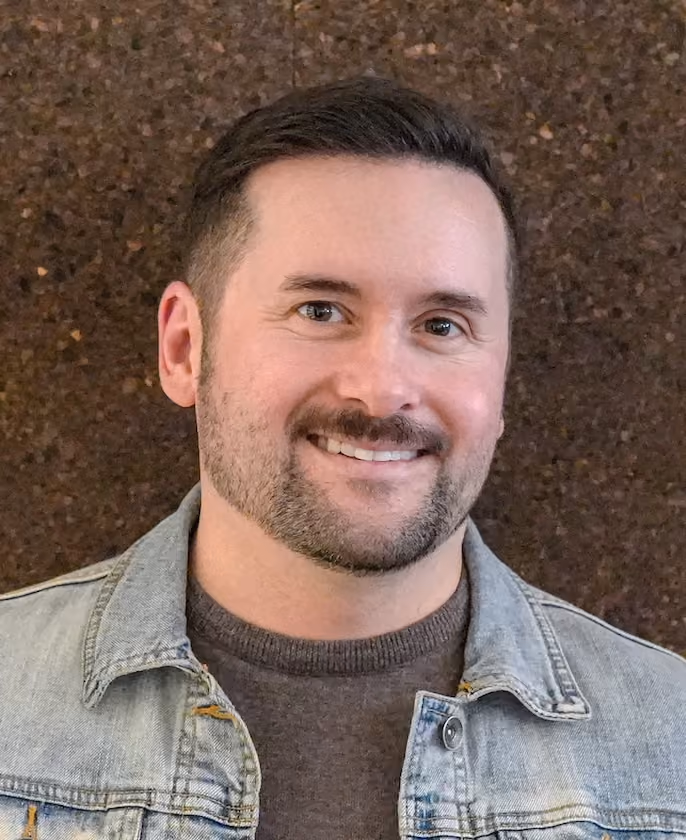 Smiling man with short dark hair and trimmed beard wearing a denim jacket over a dark sweater, standing against a brown textured background.