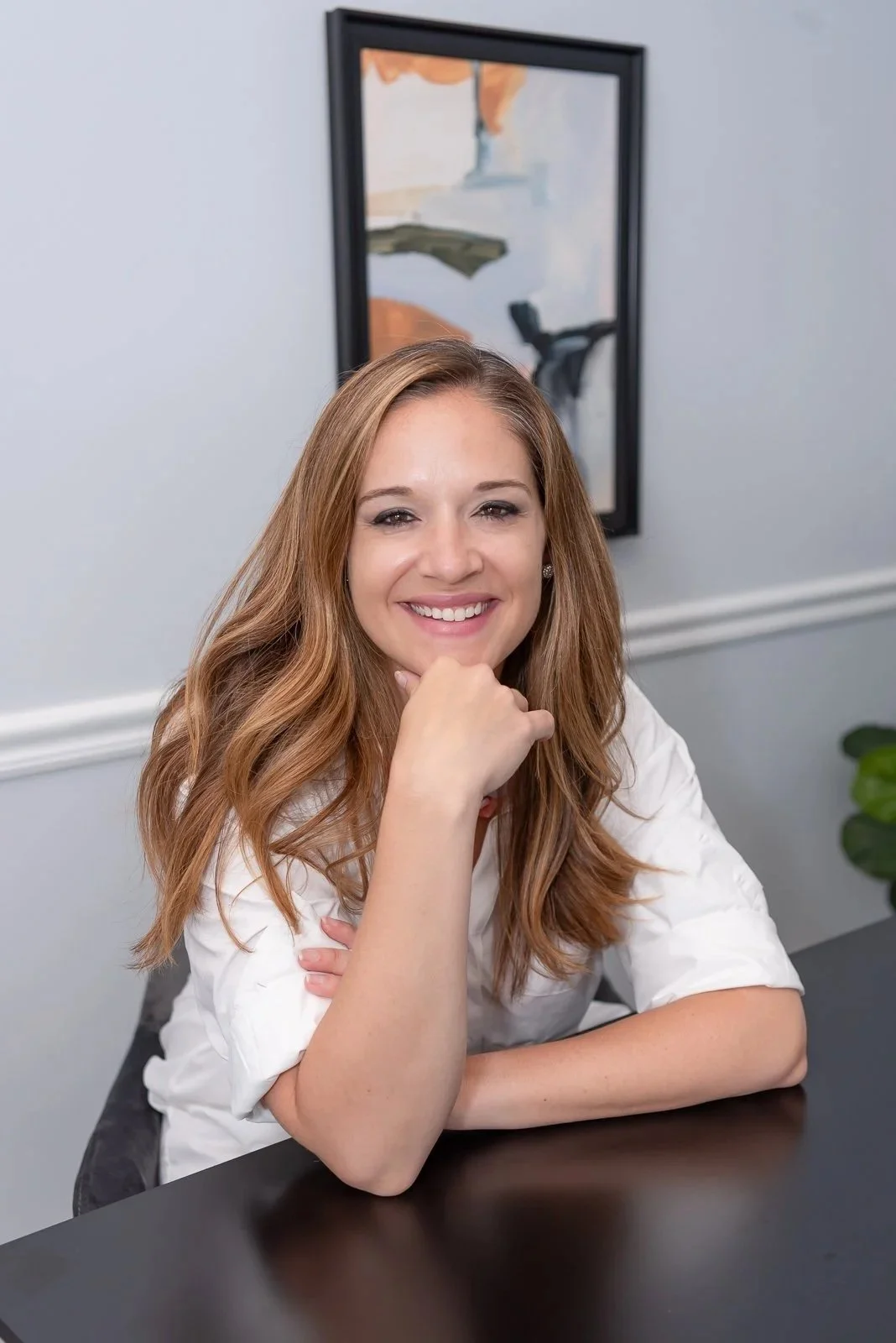 Smiling woman with long light brown hair sitting at a black table, resting her chin on her hand, with an abstract painting on the wall behind her.