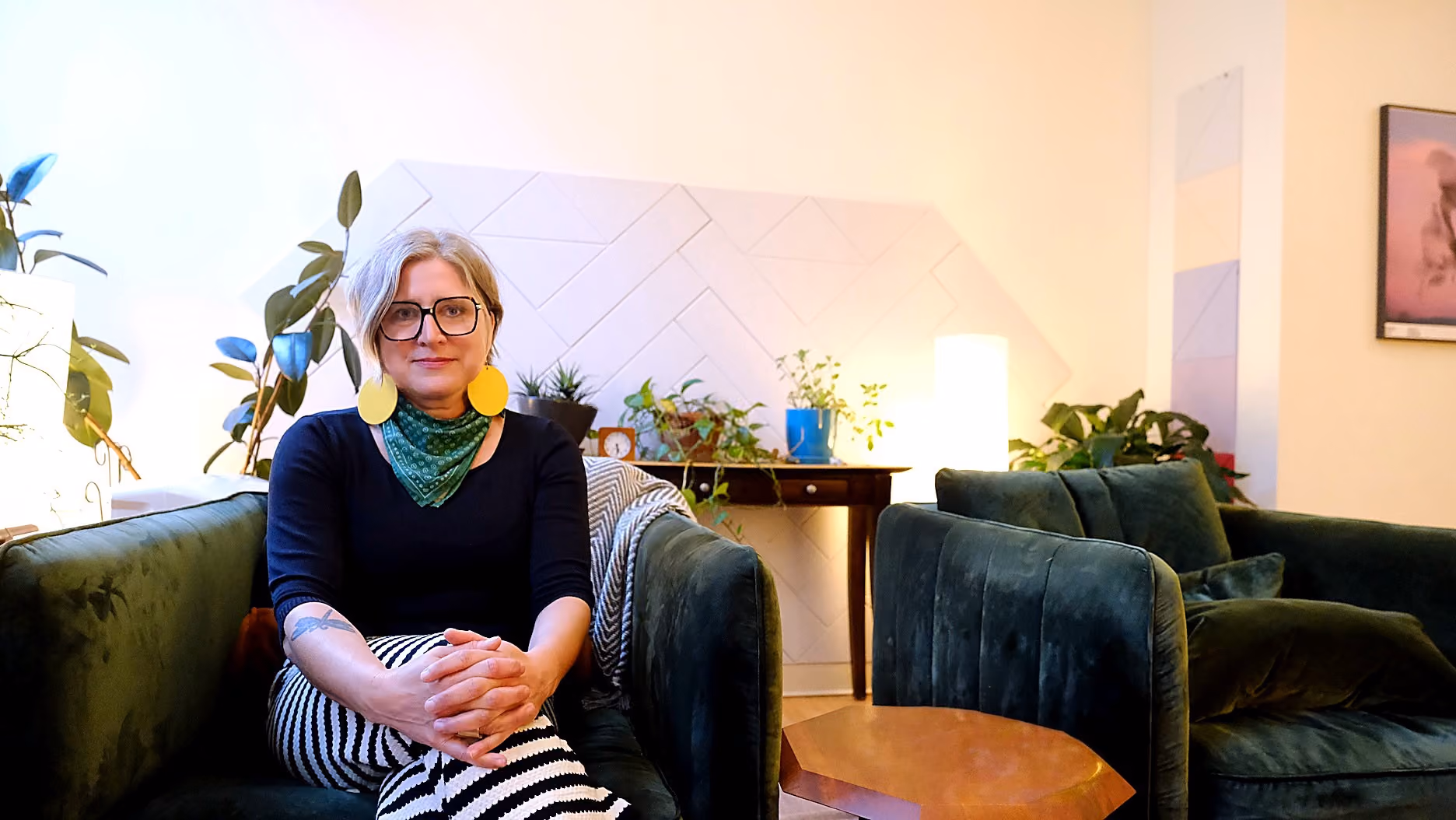 Person with short blonde hair, glasses, large yellow earrings, and a green scarf sitting on a dark green velvet couch, hands clasped, in a cozy room with plants, a wooden table, and warm lighting.