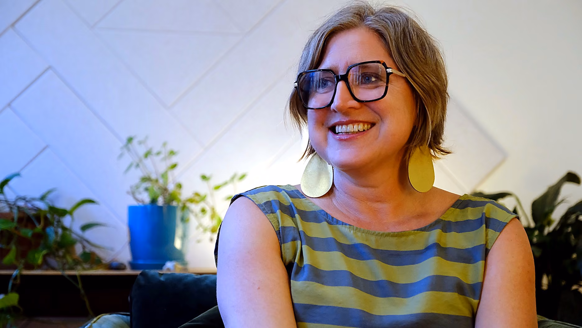 Smiling woman with short light brown hair, large glasses, and large yellow earrings wearing a green and blue striped sleeveless top sitting indoors with plants and white wall panels in the background.