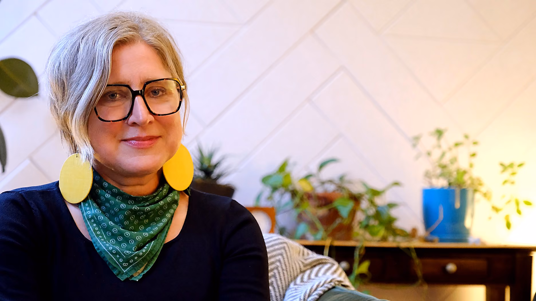 Smiling woman with short gray hair wearing large black-framed glasses, yellow earrings, and a green patterned scarf, sitting indoors with plants in the background.