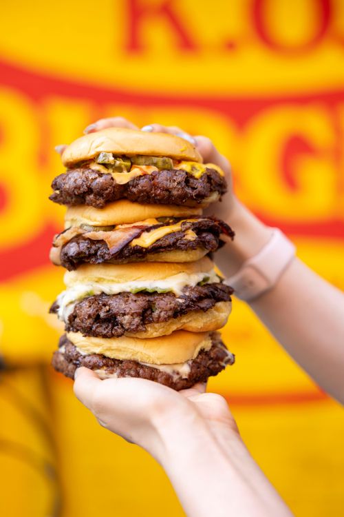 Person holding a stack of four cheeseburgers with beef patties, cheese, pickles, and bacon against a yellow and red background.