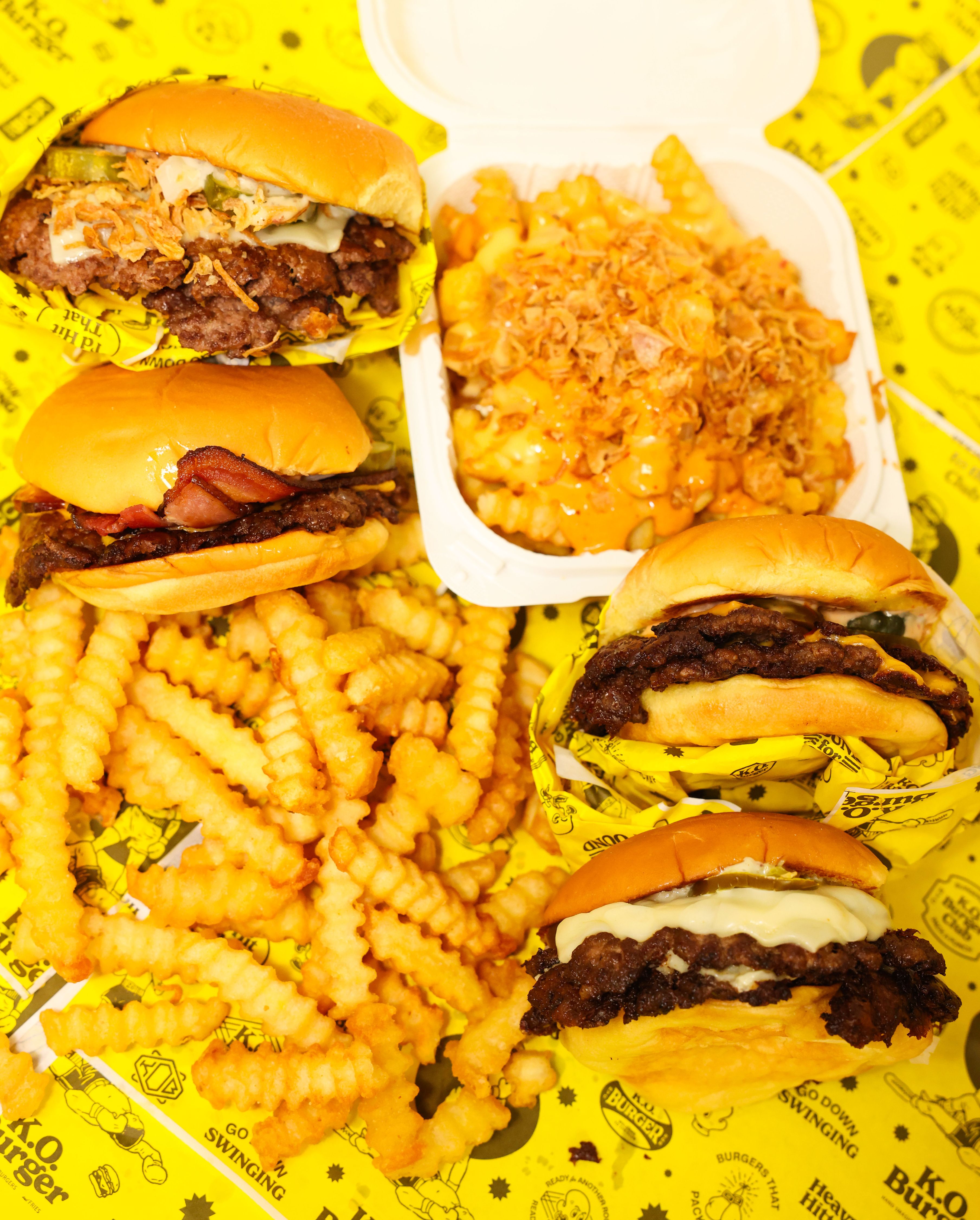 Four cheeseburgers with toppings, a serving of crinkle-cut fries, and loaded fries topped with cheese sauce and fried onions on yellow branded paper.